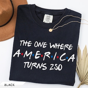May include: A black short-sleeved t-shirt featuring the white text "THE ONE WHERE AMERICA TURNS 250" with red and blue dots between the letters of the word "AMERICA". The shirt has a crew neckline and a Comfort Colors brand label at the neck.
