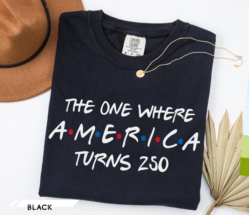 May include: A black short-sleeved t-shirt featuring the white text "THE ONE WHERE AMERICA TURNS 250" with red and blue dots between the letters of the word "AMERICA". The shirt has a crew neckline and a Comfort Colors brand label at the neck.