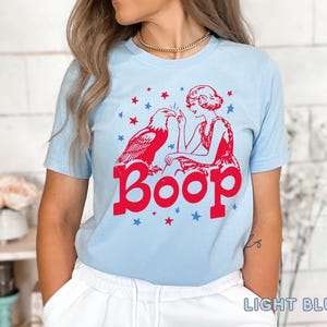 May include: Light blue t-shirt featuring a red graphic of an eagle and a woman with the word "Boop" in red. The design includes red and blue stars. The shirt is a light blue color.