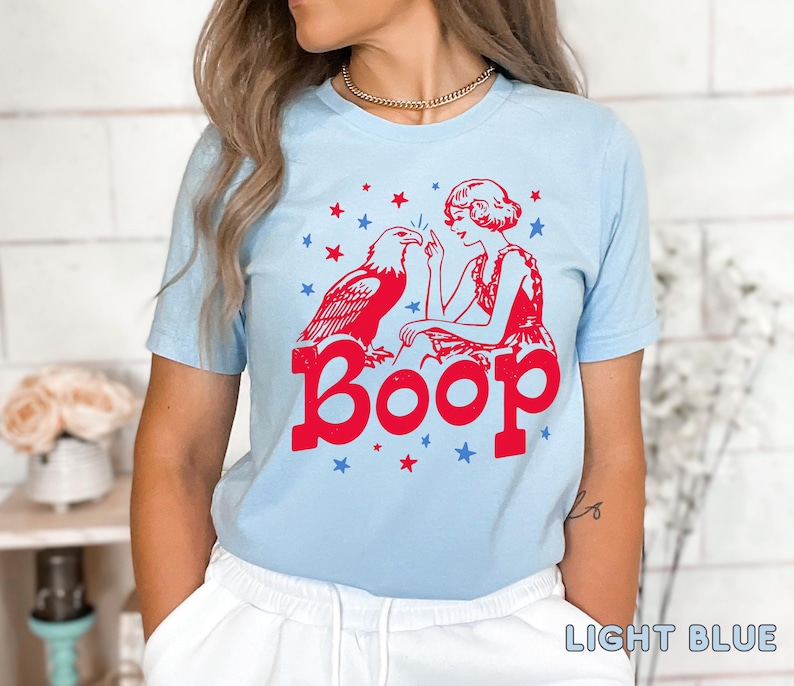May include: Light blue t-shirt featuring a red graphic of an eagle and a woman with the word "Boop" in red. The design includes red and blue stars. The shirt is a light blue color.