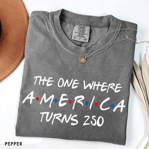 May include: A charcoal gray short-sleeve t-shirt with a crew neckline. The shirt features white text that reads THE ONE WHERE AMERICA TURNS 250, with red and blue dots separating the letters in AMERICA. A Comfort Colors brand tag is visible at the neck.