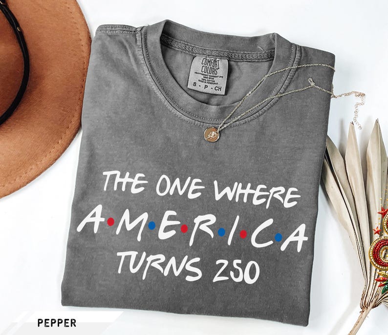 May include: A charcoal gray short-sleeve t-shirt with a crew neckline. The shirt features white text that reads THE ONE WHERE AMERICA TURNS 250, with red and blue dots separating the letters in AMERICA. A Comfort Colors brand tag is visible at the neck.