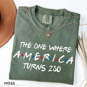 May include: A moss green short-sleeve t-shirt with a crew neckline. The shirt features white text that reads THE ONE WHERE AMERICA TURNS 250, with red and blue dots between the letters of AMERICA. A gold-toned pendant necklace rests on the fabric.