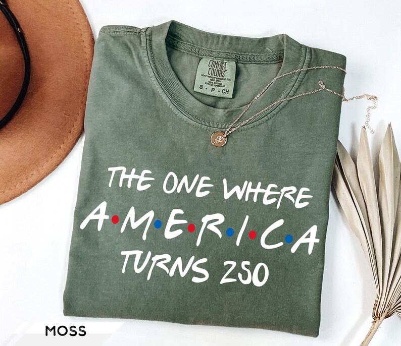 May include: A moss green short-sleeve t-shirt with a crew neckline. The shirt features white text that reads THE ONE WHERE AMERICA TURNS 250, with red and blue dots between the letters of AMERICA. A gold-toned pendant necklace rests on the fabric.