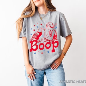 May include: Athletic heather gray t-shirt with a red graphic of an eagle, a woman, and the word "Boop." The design includes red and blue stars. The shirt is made of a soft material.