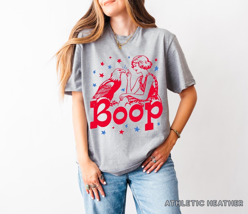 May include: Athletic heather gray t-shirt with a red graphic of an eagle, a woman, and the word "Boop." The design includes red and blue stars. The shirt is made of a soft material.