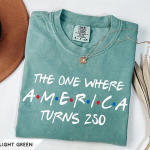 May include: A light green short-sleeve t-shirt with white text that reads THE ONE WHERE AMERICA TURNS 250, featuring red and blue dots between the letters of AMERICA. The shirt has a Comfort Colors brand label on the inside of the collar.