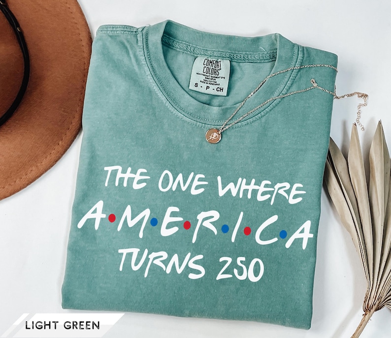 May include: A light green short-sleeve t-shirt with white text that reads THE ONE WHERE AMERICA TURNS 250, featuring red and blue dots between the letters of AMERICA. The shirt has a Comfort Colors brand label on the inside of the collar.