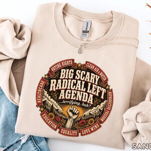 May include: A sand-colored crewneck sweatshirt featuring a graphic print with the text BIG SCARY RADICAL LEFT AGENDA ...terrifying, huh? The design includes a raised fist, mountains, flowers, and banners labeled voting rights, clean air & water, reproductive freedom, love wins, equality, education, healthcare.