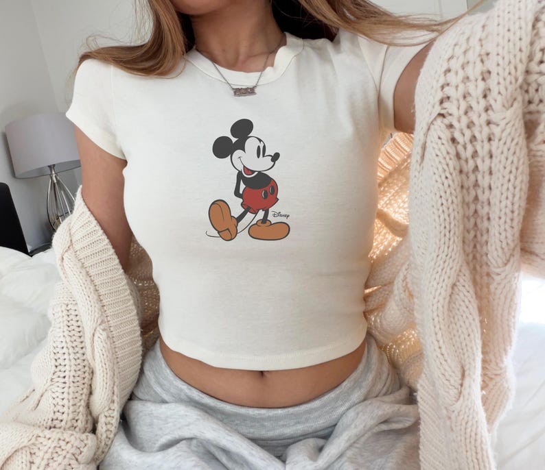 May include: Cream-colored crop top featuring a classic Mickey Mouse graphic. The cartoon character is depicted in black, red, and tan, with the Disney logo below. The top has short sleeves and a rounded neckline.