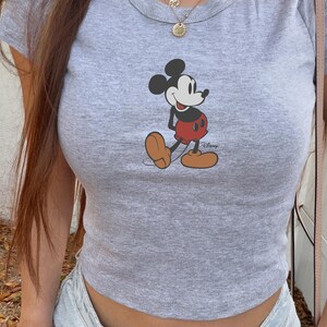 May include: A gray, short-sleeved crop top featuring a graphic of Mickey Mouse. The cartoon character is depicted in his classic outfit: red shorts, brown shoes, and black gloves. The shirt has a crew neck and is made of a soft, casual fabric.