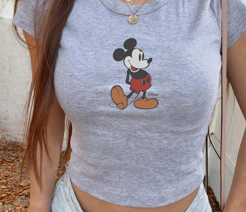 May include: A gray, short-sleeved crop top featuring a graphic of Mickey Mouse. The cartoon character is depicted in his classic outfit: red shorts, brown shoes, and black gloves. The shirt has a crew neck and is made of a soft, casual fabric.