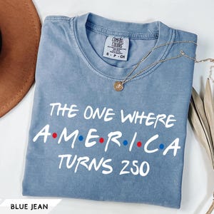 May include: A blue jean colored short-sleeve t-shirt with a crew neckline. The shirt features white text that reads THE ONE WHERE A.M.E.R.I.C.A TURNS 250, with red and blue dots separating the letters in the word AMERICA. A small gold necklace rests on the shirt.