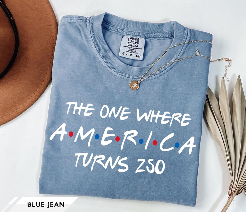 May include: A blue jean colored short-sleeve t-shirt with a crew neckline. The shirt features white text that reads THE ONE WHERE A.M.E.R.I.C.A TURNS 250, with red and blue dots separating the letters in the word AMERICA. A small gold necklace rests on the shirt.