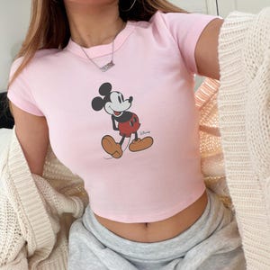 May include: A light pink crop top featuring a classic Mickey Mouse graphic. The cartoon character is depicted in black, red, and tan colors. The Disney logo is visible on the bottom right of the graphic.