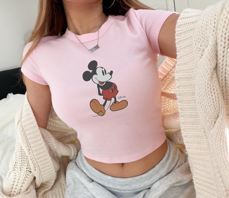 May include: A light pink crop top featuring a classic Mickey Mouse graphic. The cartoon character is depicted in black, red, and tan colors. The Disney logo is visible on the bottom right of the graphic.