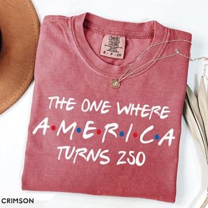 May include: A crimson short-sleeve t-shirt with a crew neckline. The shirt features white text that reads THE ONE WHERE A-M-E-R-I-C-A TURNS 250, with small red and blue dots between the letters of the word AMERICA. The shirt has a size small label.