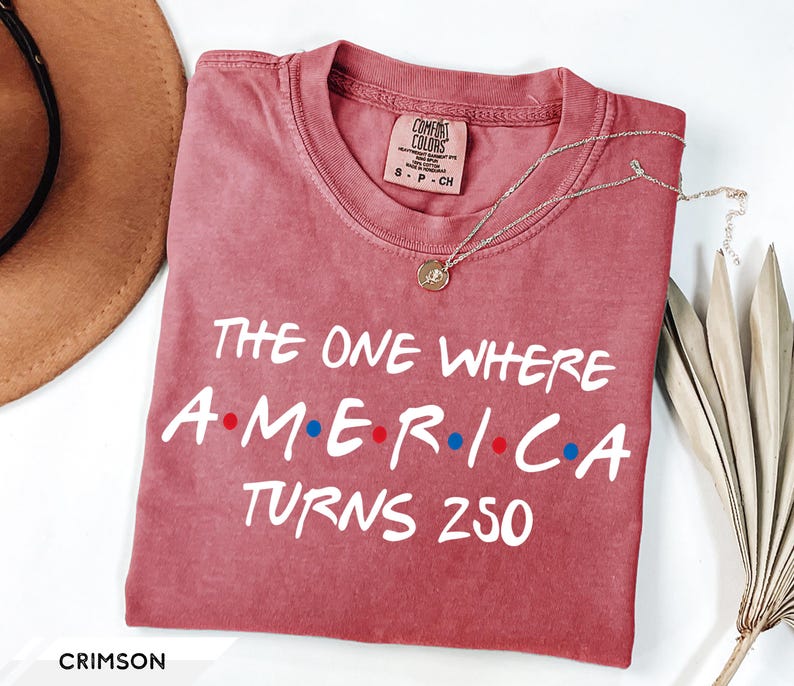 May include: A crimson short-sleeve t-shirt with a crew neckline. The shirt features white text that reads THE ONE WHERE A-M-E-R-I-C-A TURNS 250, with small red and blue dots between the letters of the word AMERICA. The shirt has a size small label.