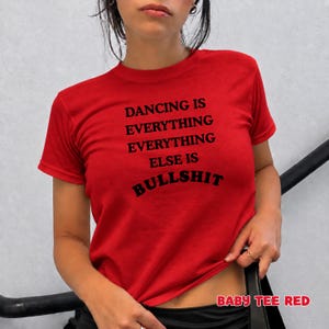 May include: A bright red short-sleeved t-shirt featuring black text printed on the front that reads DANCING IS EVERYTHING EVERYTHING ELSE IS BULLSHIT. The garment has a classic crew neckline and a straight hemline, providing a simple fit.