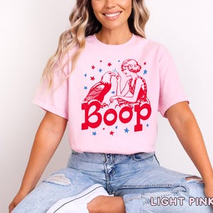 May include: Light pink t-shirt with a red graphic featuring an eagle, a woman, and the word "Boop." Blue stars are scattered around the design. The shirt is a casual style.