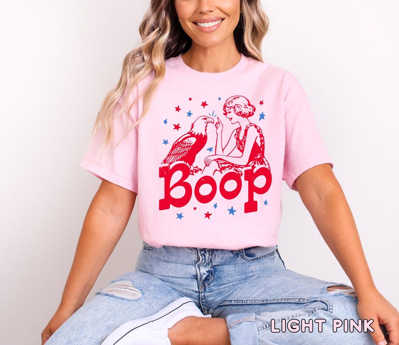 May include: Light pink t-shirt with a red graphic featuring an eagle, a woman, and the word "Boop." Blue stars are scattered around the design. The shirt is a casual style.
