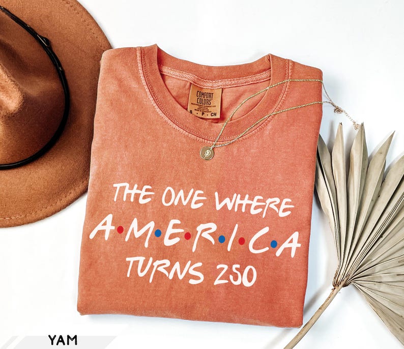 May include: A terracotta-colored short-sleeve t-shirt featuring the white text "THE ONE WHERE A.M.E.R.I.C.A TURNS 250" with red and blue dots between the letters. The shirt has a crew neckline and a Comfort Colors brand label at the inner collar.