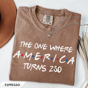 May include: An espresso-colored short-sleeve t-shirt with a crew neckline. The shirt features white text that reads THE ONE WHERE A dot M dot E dot R dot I dot C dot A TURNS 250, with red and blue dots between the letters. The label reads Comfort Colors.