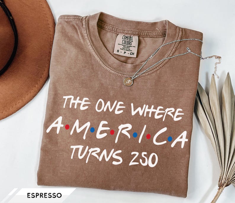 May include: An espresso-colored short-sleeve t-shirt with a crew neckline. The shirt features white text that reads THE ONE WHERE A dot M dot E dot R dot I dot C dot A TURNS 250, with red and blue dots between the letters. The label reads Comfort Colors.