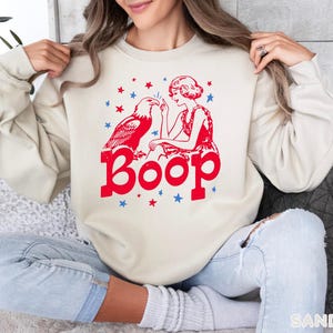 May include: A sand-colored sweatshirt with a red and blue graphic. The graphic features an eagle, a woman, stars, and the word "Boop" in bold letters. The sweatshirt is a casual style.