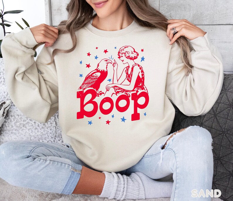 May include: A sand-colored sweatshirt with a red and blue graphic. The graphic features an eagle, a woman, stars, and the word "Boop" in bold letters. The sweatshirt is a casual style.