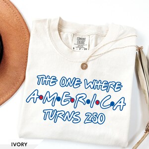 May include: An ivory short-sleeve t-shirt with a crew neckline featuring blue text that reads The One Where A-M-E-R-I-C-A Turns 250, with small red and blue dots between the letters of the word America. The shirt has a Comfort Colors brand label.