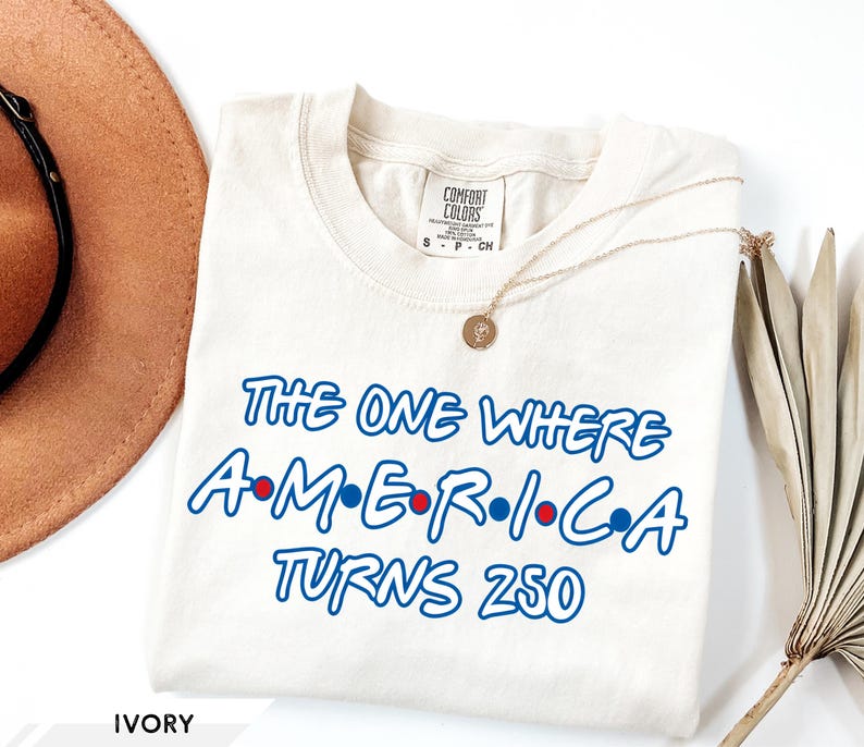 May include: An ivory short-sleeve t-shirt with a crew neckline featuring blue text that reads The One Where A-M-E-R-I-C-A Turns 250, with small red and blue dots between the letters of the word America. The shirt has a Comfort Colors brand label.
