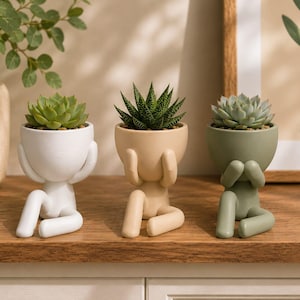 Può includere: Tre piccoli vasi in ceramica a forma di figure sedute con le mani sulla testa. Ogni vaso contiene una diversa pianta succulenta. Le figure sono di colore bianco opaco, beige chiaro e verde salvia, disposte in fila su una superficie.