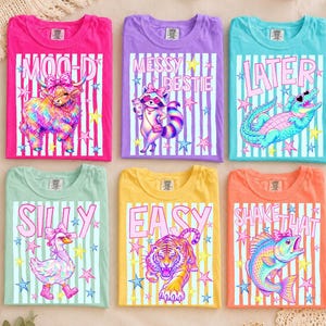 Puede incluir: Seis camisetas gráficas coloridas. Cada camiseta presenta un diseño diferente: una vaca de las Highlands, un mapache, un caimán, un ganso, un tigre y un pez. Las camisetas tienen el texto "MOOD", "MESSY BESTIE", "LATER", "SILLY", "EASY" y "SHAKE THAT".