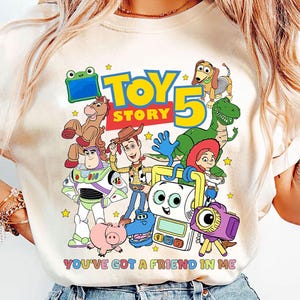 May include: A cream-colored short-sleeve t-shirt featuring a colorful graphic print of Toy Story characters including Woody, Buzz Lightyear, Jessie, Rex, and Slinky Dog. The text reads Toy Story 5 and You've Got A Friend In Me with small yellow stars.