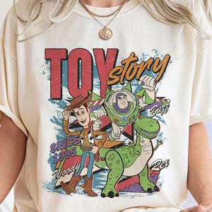May include: Cream-colored t-shirt featuring a colorful graphic of Toy Story characters Woody, Buzz Lightyear, and Rex. The design includes the text "TOY STORY" and "SINCE 1995". The shirt has short sleeves and a relaxed fit.