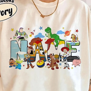 May include: Ivory-colored t-shirt with the text "NAME" in a colorful, cartoon-style design. The design features various characters and elements from a popular animated movie, including a green dinosaur, a space ship, and a cowboy.