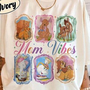 以下が含まれることがあります： 「Comfort Colors」と「Ivory」の文字が円形にデザインされたアイボリー色のTシャツ。ディズニーキャラクターのイラストがフレームに入っており、「Mom Vibes」の文字がカラフルなスクリプトで描かれています。