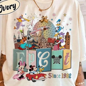 May include: An ivory short-sleeve t-shirt featuring a colorful graphic print of various cartoon characters, the Epcot geodesic sphere, and the text Epcot Since 1982. The design includes Goofy, Donald Duck, Daisy Duck, Minnie Mouse, and Pluto.