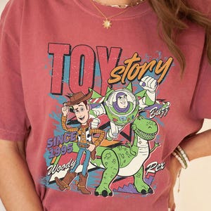 May include: A maroon t-shirt featuring a graphic of characters from the animated film "Toy Story." The design includes Woody, Buzz Lightyear, and Rex, with the text "TOY STORY" and "SINCE 1995." The shirt has short sleeves.