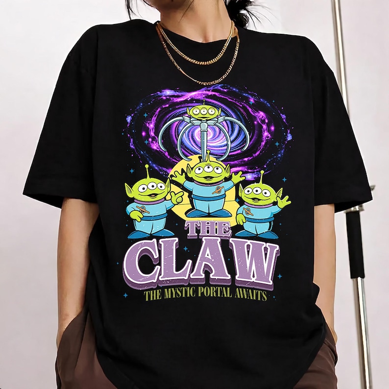 May include: A black short-sleeved t-shirt featuring a graphic of three green three-eyed aliens in blue space suits. Above them is a mechanical claw and a purple galaxy portal. The text reads THE CLAW and THE MYSTIC PORTAL AWAITS in white and purple.