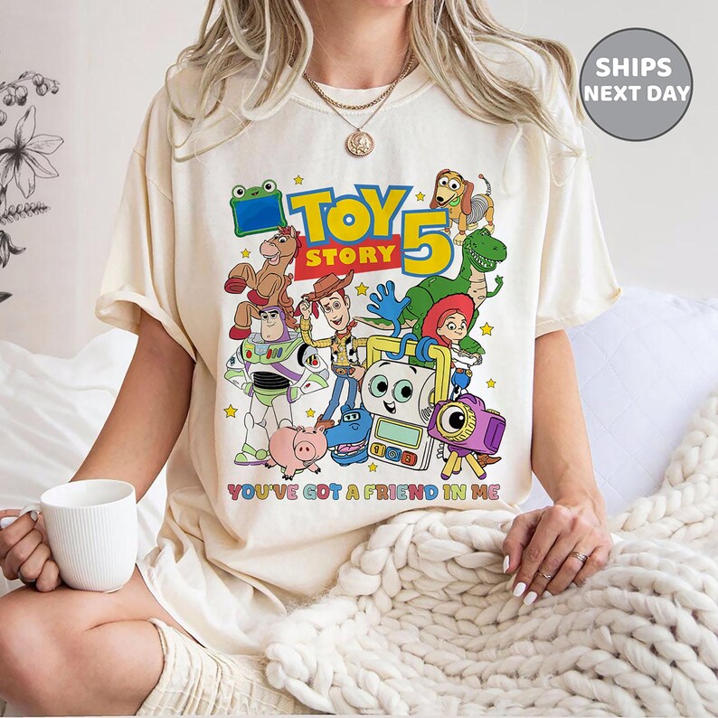May include: A cream-colored t-shirt featuring a colorful graphic of Toy Story characters including Woody, Buzz Lightyear, Rex, and Slinky Dog. The text reads "TOY STORY 5" and "YOU'VE GOT A FRIEND IN ME" in bold, multi-colored lettering across the front.