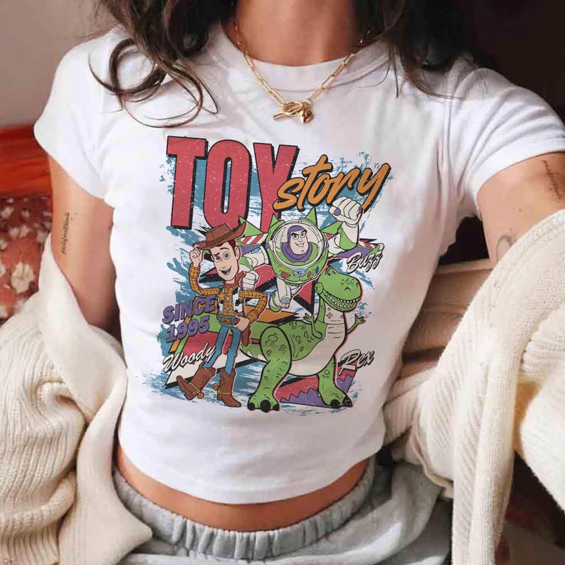 May include: White t-shirt featuring a colorful graphic of characters from the animated film "Toy Story." The design includes Woody, Buzz Lightyear, and Rex, with the words "Toy Story" and "Since 1995" prominently displayed.
