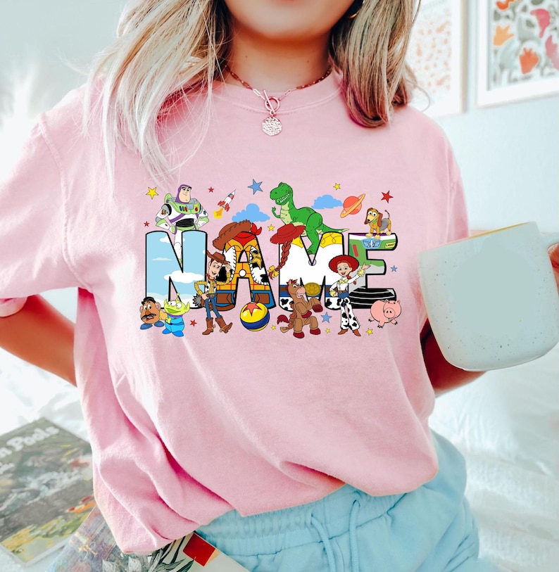 May include: Pink t-shirt featuring the word "NAME" in a colorful, cartoon-style design. The letters are filled with characters from a popular animated movie. The shirt is a light pink color.