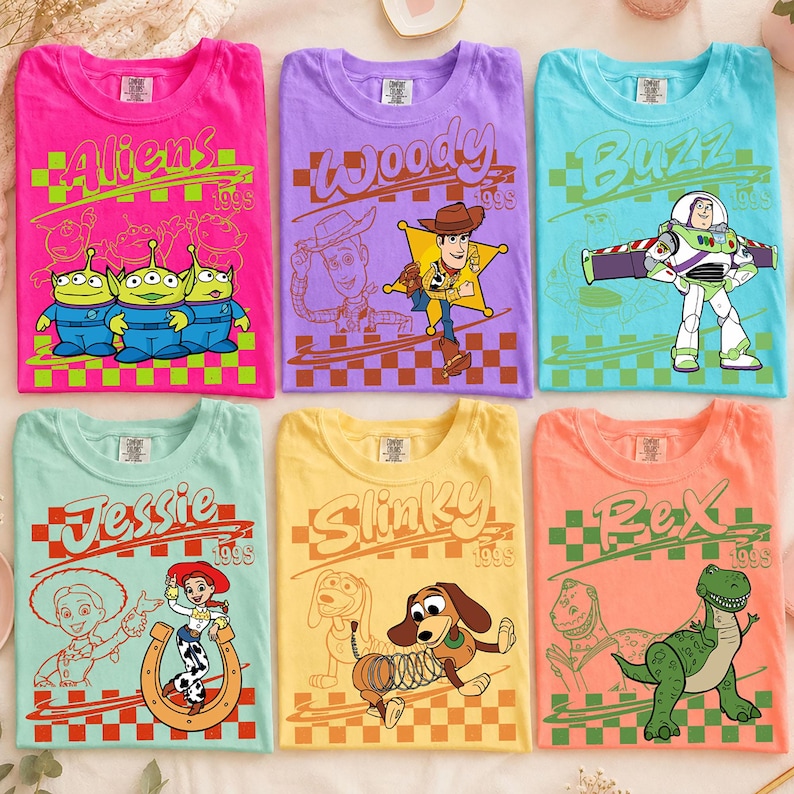 K&ouml;nnte beinhalten: Sechs kurz&auml;rmelige T-Shirts in Pink, Lila, Hellblau, Mint, Gelb und Orange. Jedes Shirt zeigt eine Toy Story-Charaktergrafik, darunter Aliens, Woody, Buzz Lightyear, Jessie, Slinky Dog und Rex, mit dem Text 1995 und einem Schachbrettmuster.