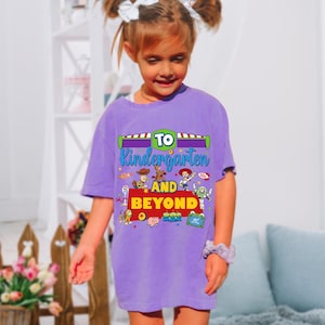 May include: A purple short-sleeve t-shirt featuring a graphic print with the text TO KINDERGARTEN AND BEYOND. The design includes colorful illustrations of various animated toy characters, including a cowboy, a space ranger, and a cowgirl.