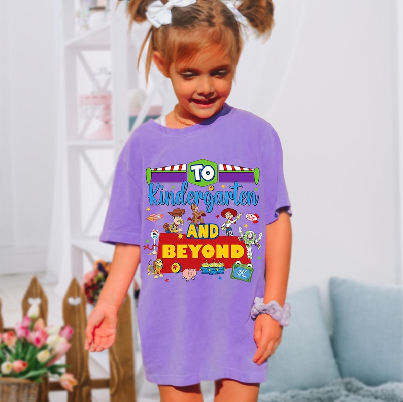 May include: A purple short-sleeve t-shirt featuring a graphic print with the text TO KINDERGARTEN AND BEYOND. The design includes colorful illustrations of various animated toy characters, including a cowboy, a space ranger, and a cowgirl.