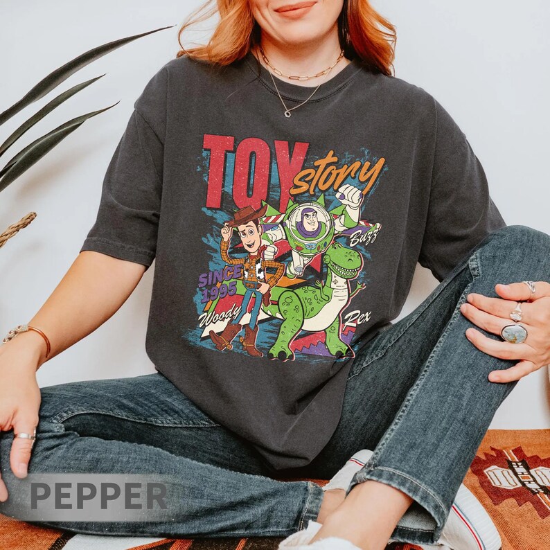 May include: Dark gray t-shirt featuring a graphic of Toy Story characters Woody, Buzz Lightyear, and Rex. The text "TOY STORY" is in red and yellow, with "SINCE 1995" below. The shirt is a vintage style, with a slightly faded appearance.