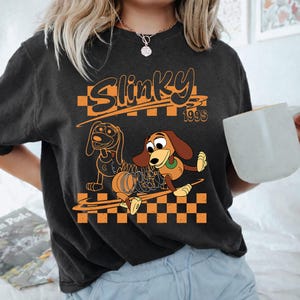 K&ouml;nnte beinhalten: Ein anthrazitfarbenes Kurzarm-T-Shirt mit einem Grafikdruck und dem Text Slinky 1990s. Das Design zeigt zwei Illustrationen eines braunen Dackel-Spielzeugs mit einem Metallfederk&ouml;rper vor einem orange-schwarzen Schachbrettmuster.