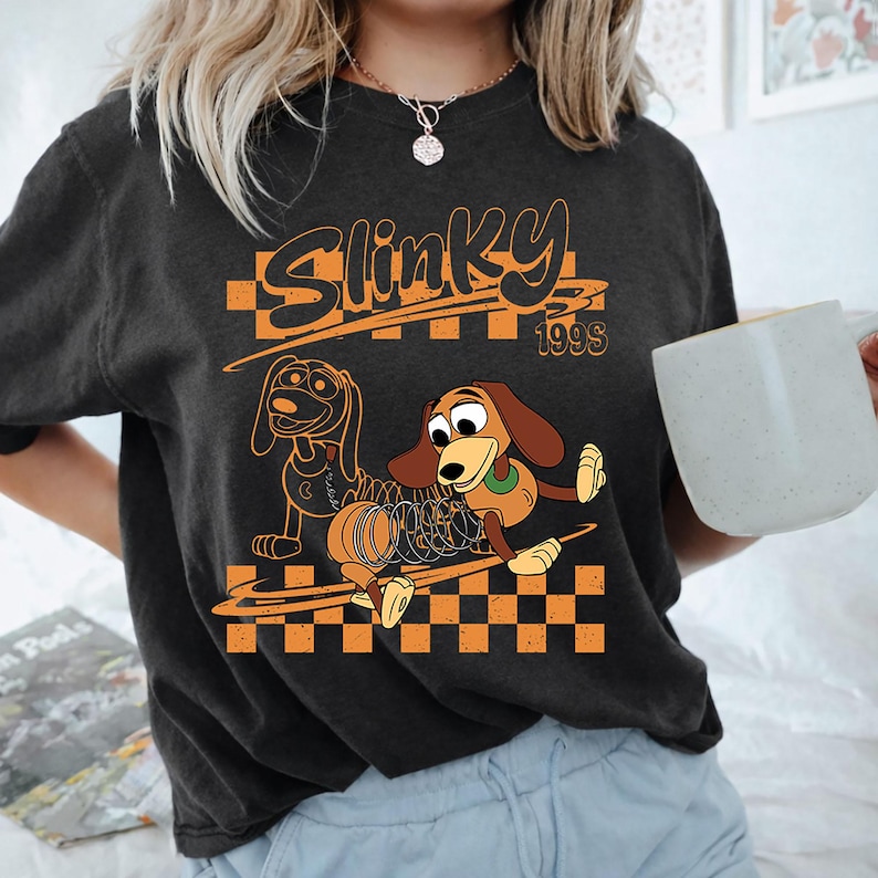 K&ouml;nnte beinhalten: Ein anthrazitfarbenes Kurzarm-T-Shirt mit einem Grafikdruck und dem Text Slinky 1990s. Das Design zeigt zwei Illustrationen eines braunen Dackel-Spielzeugs mit einem Metallfederk&ouml;rper vor einem orange-schwarzen Schachbrettmuster.